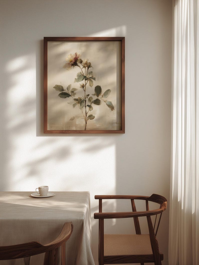Puede incluir: Una impresi&oacute;n bot&aacute;nica enmarcada con una rama de rosa con hojas verdes y flores rosas y amarillas. La obra de arte est&aacute; en un marco de madera oscura, colgada en una pared blanca. Una mesa con un mantel neutro y una taza y platillo est&aacute;n en primer plano.