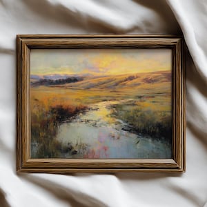 May include: A framed landscape painting depicts a serene scene with a river flowing through a field. The artwork features warm tones of yellow, orange, and brown, with hints of blue and green. The painting is encased in a brown wooden frame.