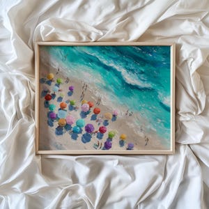 May include: A framed print of a beach scene with colourful parasols and turquoise waves. The painting shows people on the sand and the ocean. The artwork is in a light wooden frame.