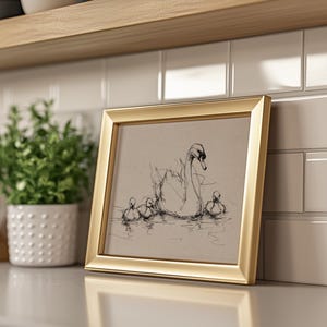 May include: A gold framed print featuring a sketch of a swan and three cygnets in a body of water. The artwork is displayed on a white countertop next to a potted plant and a white tile backsplash.