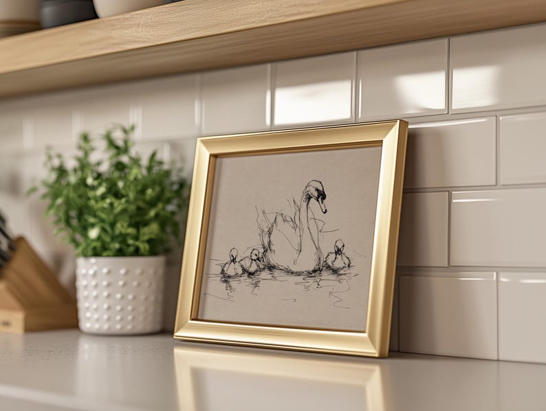 May include: A gold framed print featuring a sketch of a swan and three cygnets in a body of water. The artwork is displayed on a white countertop next to a potted plant and a white tile backsplash.
