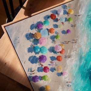 May include: A painting depicting a beach scene from above, showing colourful parasols and people on the sand. The artwork has a sandy beach, turquoise water, and a wooden frame. The parasols are in shades of red, pink, and yellow.