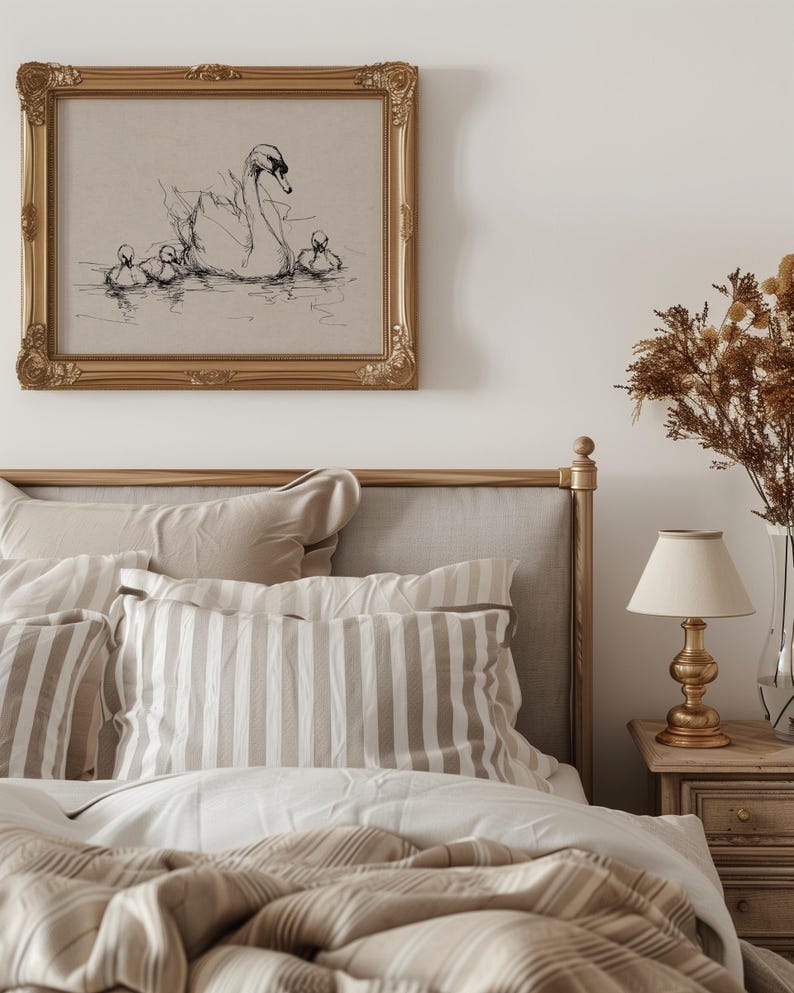 May include: A bedroom scene featuring a bed with beige and white striped pillows and bedding. Above the bed is a framed artwork with a sketch of a swan and cygnets. A gold-toned lamp sits on a wooden nightstand.