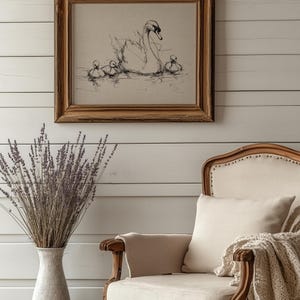 May include: A framed pen and ink drawing of a swan with cygnets on water. The artwork is in a wooden frame and hangs on a white paneled wall. A vase of lavender and a cream-colored armchair are in the foreground.