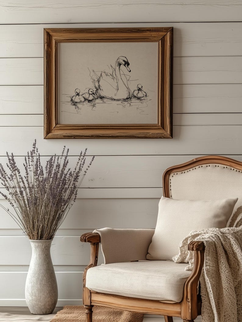 May include: A framed pen and ink drawing of a swan with cygnets on water. The artwork is in a wooden frame and hangs on a white paneled wall. A vase of lavender and a cream-colored armchair are in the foreground.