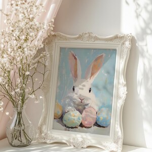 May include: A white rabbit with long ears sits surrounded by colorful Easter eggs in a white frame. The frame is on a white shelf with a vase of white flowers in the background.