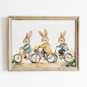 May include: Three watercolor bunnies riding bicycles. The bunnies are wearing blue and yellow clothing. The bicycles are red and have baskets. The background is white.