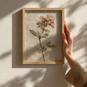 May include: A framed floral print featuring a delicate botanical illustration. The artwork showcases a sprig of flowers in soft pink and peach hues, with green leaves against a neutral background. The frame is made of light-colored wood.