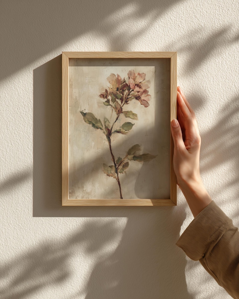 May include: A framed floral print featuring a delicate botanical illustration. The artwork showcases a sprig of flowers in soft pink and peach hues, with green leaves against a neutral background. The frame is made of light-colored wood.