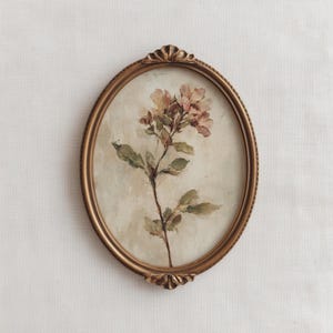 May include: An oval-shaped, antique-style framed floral painting. The artwork features a delicate sprig of pink and green flowers against a neutral background. The frame is a detailed gold color, adding to the vintage aesthetic.