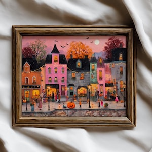 Op de afbeelding: Een ingelijste kunstdruk met een stadsstraatscène bij schemering met roze en oranje luchten. De illustratie toont veelkleurige gebouwen, verlichte straatlantaarns, lichtsnoeren en mensen die op een kasseienpad lopen met een grote oranje pompoen op de voorgrond.