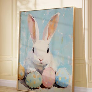 May include: A white rabbit with pink ears sits surrounded by four colorful Easter eggs. The eggs are painted in shades of blue, yellow, pink, and green. The background is a light blue with a white speckled pattern.