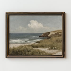 May include: A framed landscape painting depicts a coastal scene. The artwork features a sandy beach, ocean waves, and a grassy cliff under a cloudy sky. The frame is dark brown with a gold inner edge, enhancing the vintage aesthetic.
