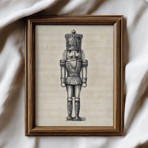 Puede incluir: Una impresión enmarcada en blanco y negro de un soldado cascanueces. La obra de arte presenta una ilustración detallada de la figura tradicional, sobre un fondo de partituras. El marco es de color marrón cálido.