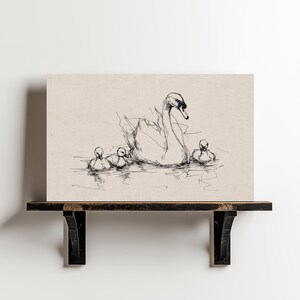 May include: A black and white sketch of a swan and its cygnets swimming in water. The artwork is on a beige background, creating a minimalist and elegant aesthetic. The drawing is displayed on a dark wooden shelf against a white wall.