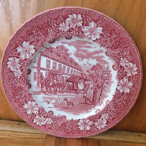 May include: A round, antique plate with a red and white floral border. The center features a detailed scene of a building with horses and people. The plate has a scalloped edge and is likely made of ceramic.