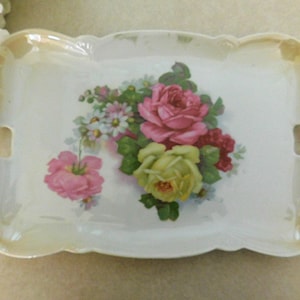 May include: A rectangular, white porcelain serving tray with a scalloped edge and two handles. The tray features a floral design of pink, red, and yellow roses with green leaves. The rim has a gold-colored trim.