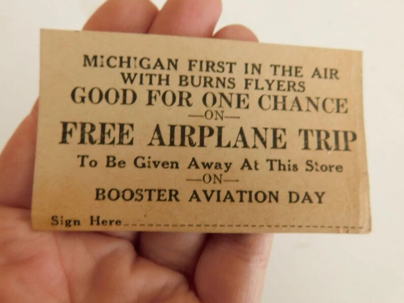 Puede incluir: Una tarjeta vintage de color beige con texto negro. La tarjeta dice "MICHIGAN FIRST IN THE AIR WITH BURNS FLYERS GOOD FOR ONE CHANCE ON FREE AIRPLANE TRIP." La tarjeta es para el Booster Aviation Day.