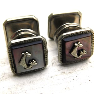 May include: A pair of vintage cufflinks with a square face featuring a mother-of-pearl inlay. Each cufflink has a silver-toned frame with a decorative edge and a small anchor emblem. The cufflinks are set against a white background.