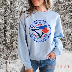 Puede incluir: Sudadera azul claro con el logotipo de los Toronto Blue Jays. El logotipo es circular y presenta un arrendajo azul, una hoja de arce roja y las palabras "Toronto Blue Jays". El texto "LIGHT BLUE" está en la parte inferior.