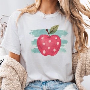 May include: A white short-sleeved t-shirt featuring a graphic of a red apple with white polka dots and a green leaf. The apple is centered over a horizontal teal brushstroke design. The shirt has a crew neckline and a standard fit.