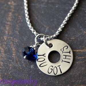 May include: A silver necklace with a round charm that says "You got this". The charm has a hole in the center and is attached to a silver chain. A small blue crystal is attached to the chain above the charm.
