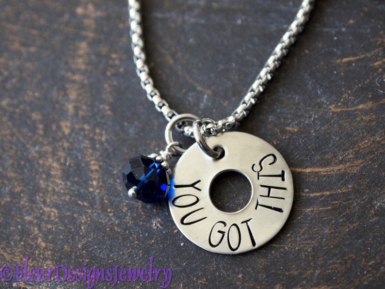 May include: A silver necklace with a round charm that says "You got this". The charm has a hole in the center and is attached to a silver chain. A small blue crystal is attached to the chain above the charm.