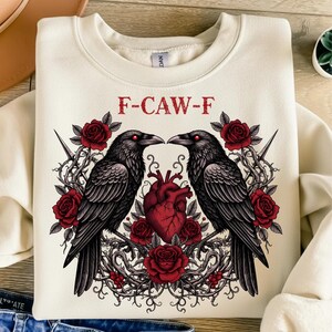 Puede incluir: Sudadera crema con un dise&ntilde;o g&oacute;tico. Presenta dos cuervos negros enfrentados, rosas rojas, un coraz&oacute;n rojo y el texto "F-CAW-F" en rojo. El dise&ntilde;o es detallado e intrincado.