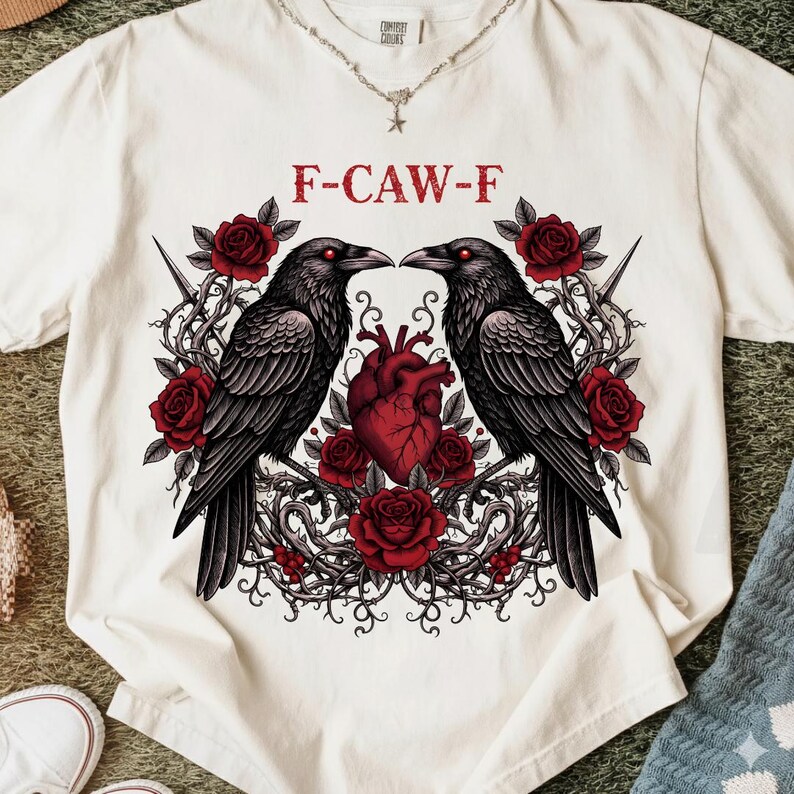 Puede incluir: Camiseta color crema con un dise&ntilde;o g&oacute;tico. Dos cuervos negros se enfrentan sobre un coraz&oacute;n rojo, rodeados de rosas rojas y vides espinosas. El texto "F-CAW-F" est&aacute; impreso encima de los cuervos.