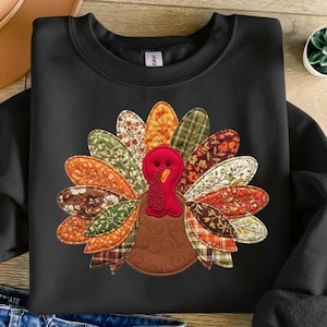 May include: Black sweatshirt featuring a colorful turkey design. The turkey has a brown body, a red head and wattle, and a fan of patterned feathers in shades of orange, yellow, green, and brown. A festive garment for Thanksgiving.
