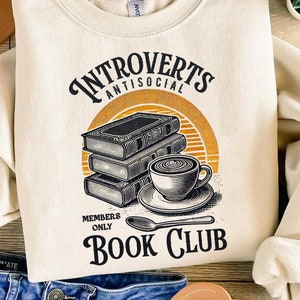 May include: Cream-coloured sweatshirt featuring the text "Introverts Antisocial Book Club". The design includes a stack of books, a coffee cup, and a spoon, in black and orange, set against a retro-style background.