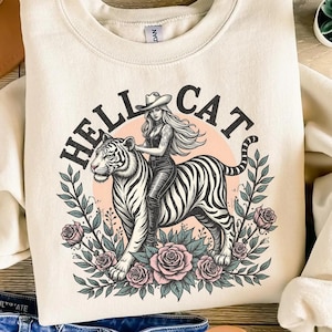 Könnte beinhalten: Ein cremefarbener Pullover mit einem Grafikdruck einer Frau, die auf einem weißen Tiger reitet. Die Grafik enthält den Text "HELLCAT" in einem schwarzen Kreisrahmen mit floralen Akzenten.