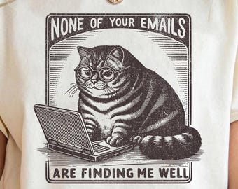 May include: A cream-colored t-shirt with a vintage-style illustration of a cat wearing glasses and using a laptop. The text on the shirt reads "None of your emails are finding me well."