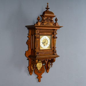 Könnte beinhalten: Eine hölzerne Wanduhr mit aufwendigen Schnitzereien und einem dekorativen Pendel. Das Zifferblatt ist weiß mit schwarzen römischen Ziffern und goldenen Akzenten. Die Uhr ist an einer grauen Wand befestigt.