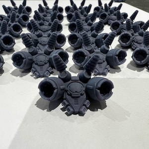 Puede incluir: Un grupo de 18 pequeñas figuras de robots impresas en 3D de color gris oscuro con múltiples protuberancias redondeadas en cada una. Las figuras están dispuestas en un patrón simétrico sobre una superficie blanca.