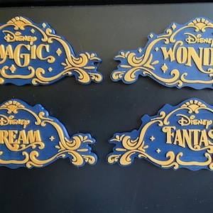 May include: Four decorative plaques featuring a dark blue base with raised gold lettering and ornate scrollwork. Each plaque displays the word Disney above the words Magic, Wonder, Dream, or Fantasy, respectively, in a stylized, elegant font.