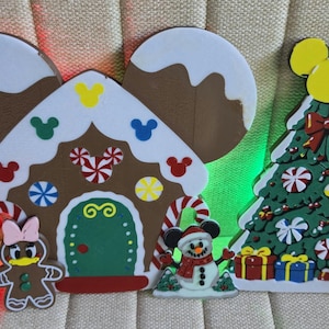 May include: A wooden gingerbread house cutout with Mickey-shaped ears, a green door, and candy accents, alongside a green Christmas tree cutout topped with yellow Mickey ears, decorated with ornaments, gifts, and two gingerbread characters.