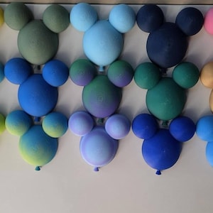 Puede incluir: Una colección de numerosos arreglos de globos con forma de cabeza de ratón, que presentan una gran variedad de colores sólidos, degradados y acabados metálicos. Cada arreglo consta de un globo central grande con dos globos más pequeños como orejas.