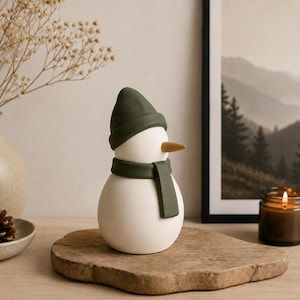 May include: A smooth, matte white snowman figurine featuring a dark olive green beanie hat and a matching scarf. The snowman has a simple, rounded body and a pointed, light tan nose protruding from the center of its head, creating a clean look.
