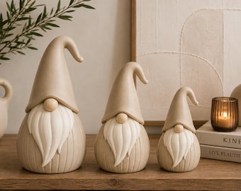 Puede incluir: Un conjunto de tres figuras de gnomos de diferentes tamaños, cada una con un sombrero puntiagudo de color beige, una nariz redonda y una barba larga, blanca y texturizada. Los cuerpos son redondeados con un patrón de rayas verticales.