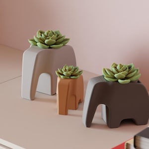 May include: Three elephant-shaped planters in varying sizes and colors, each holding a succulent plant. The planters are in shades of gray, brown, and orange. The succulents are green. The planters are on a light-colored surface.