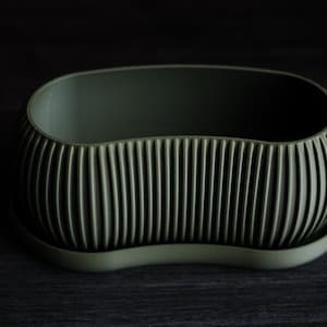 May include: An olive green planter with a ribbed exterior and a matching saucer. The oval-shaped planter is made of a matte material and sits on a dark surface. The vertical ribs add texture to the design.
