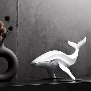 Peut inclure: Une sculpture de baleine blanche au design épuré et moderne. La baleine est posée sur une surface noire, avec un vase noir et une longue bougie noire en arrière-plan. C'est un objet décoratif.