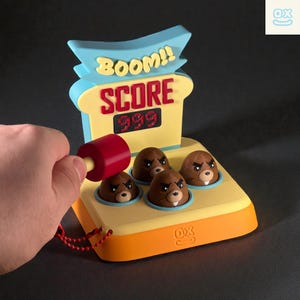 May include: A Whack-a-Mole game with a red mallet poised above a brown mole. The game features a yellow base with four mole holes, a digital score display showing "999", and the word "BOOM!!" in yellow.