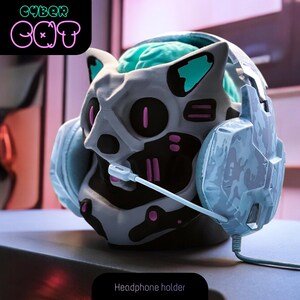 May include: A gray and black 3D printed cat head headphone holder with a blue and white camouflage headset. The cat head has pink and teal accents and the text "CYBER CAT" is in the background.
