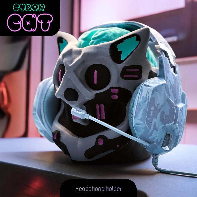May include: A gray and black 3D printed cat head headphone holder with a blue and white camouflage headset. The cat head has pink and teal accents and the text "CYBER CAT" is in the background.