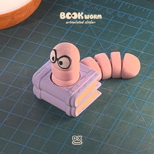Puede incluir: Un clicker articulado rosa con forma de gusano de libro. El gusano lleva gafas y está sentado sobre una pila de libros morados con páginas amarillas. El texto "BOOKWORM articulated clicker" está encima del gusano.