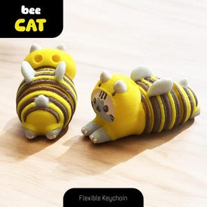 May include: Two yellow and brown bee-themed cat keychains. The keychain on the left is standing upright, while the keychain on the right is lying down. Both keychains have a flexible design and feature a bee-like pattern.