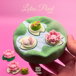 Può includere: Un pezzo decorativo rotondo, verde e bianco con il testo "Lotus Pond Clicker". Presenta un design a laghetto con tre ninfee, ognuna con una rana o un fiore di loto. Ulteriori fiori di loto e una rana sono posizionati intorno.