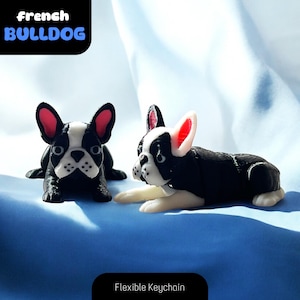Puede incluir: Dos llaveros de Bulldog Francés impresos en 3D en blanco y negro. Los llaveros son flexibles y tienen orejas rojas. Los llaveros están sobre una superficie azul.  Llavero flexible.